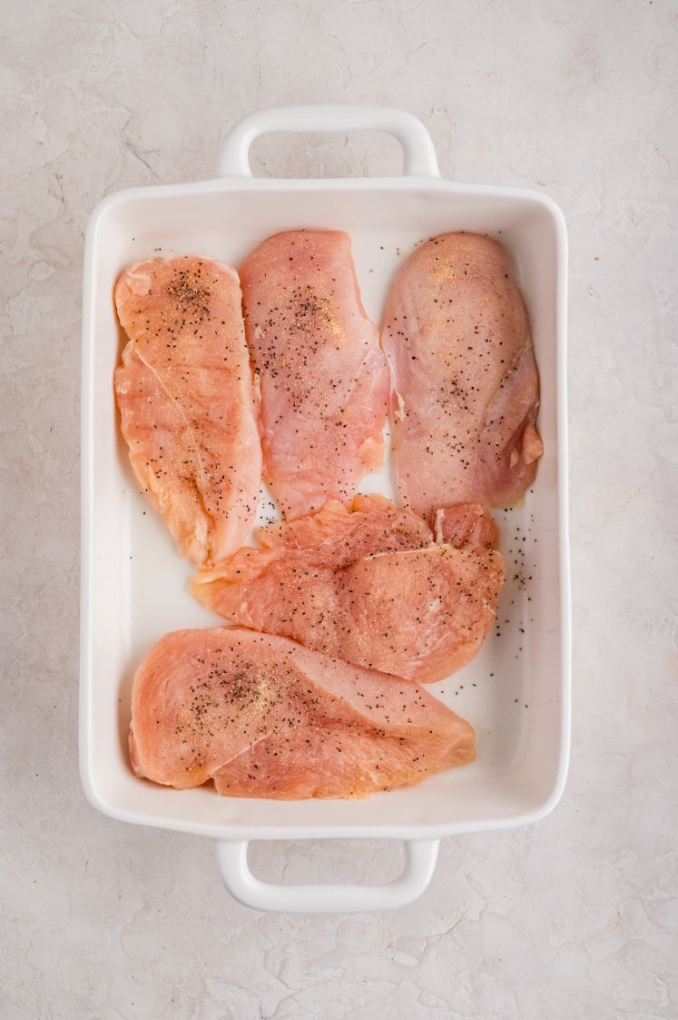Five raw chicken breasts seasoned with salt and pepper are arranged in a single layer inside a white rectangular baking dish on a light-colored surface.