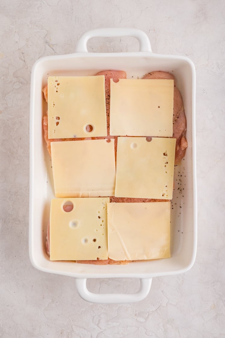 A white baking dish containing raw chicken breasts topped with six slices of Swiss cheese, arranged in two rows on a light-colored surface.