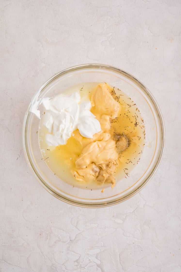 A glass bowl containing dollops of mayonnaise, sour cream, Dijon mustard, and seasonings on a light countertop, viewed from above.