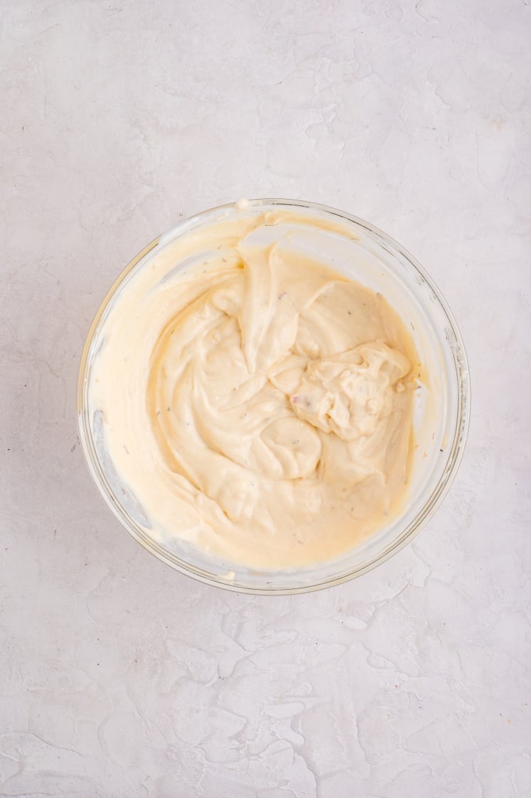A clear glass bowl filled with creamy, light beige mayonnaise sits on a textured white surface. The mayonnaise appears smooth and thick, with gentle swirls on top.