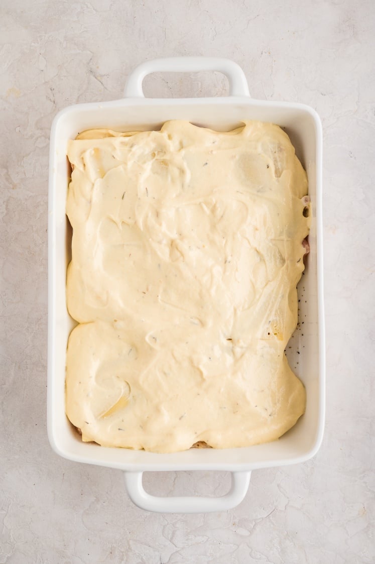 A white rectangular baking dish filled with an uncooked casserole topped with a thick, creamy sauce, placed on a light-colored textured surface.