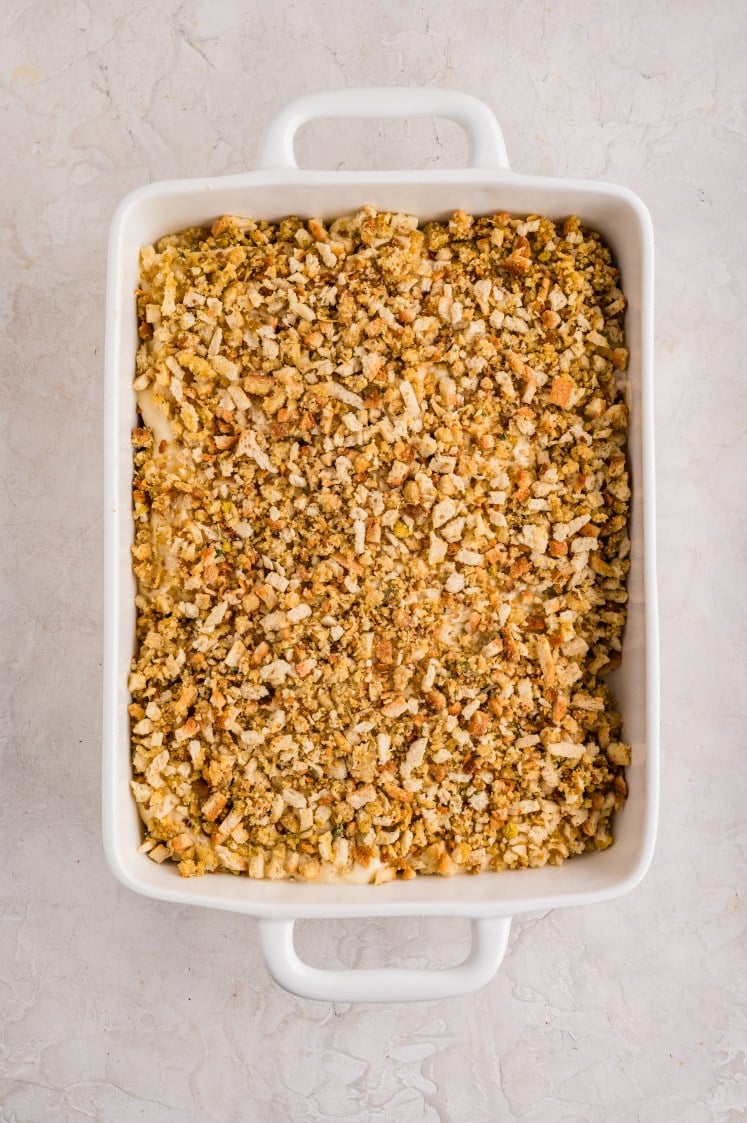 A white rectangular baking dish filled with a casserole topped with a golden-brown layer of crushed stuffing or breadcrumbs, set on a light-colored marble surface.