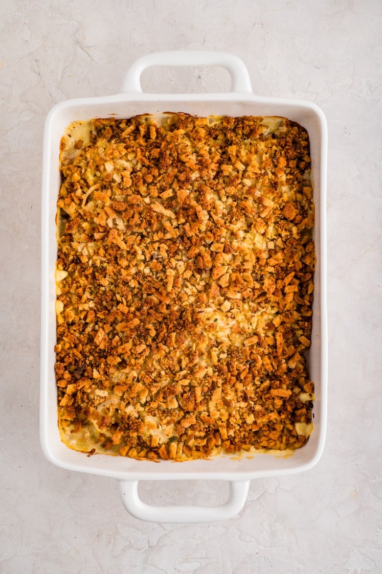A rectangular white baking dish filled with a casserole topped with a layer of golden-brown, crispy bread crumbs or stuffing. The dish sits on a light-colored, textured surface.
