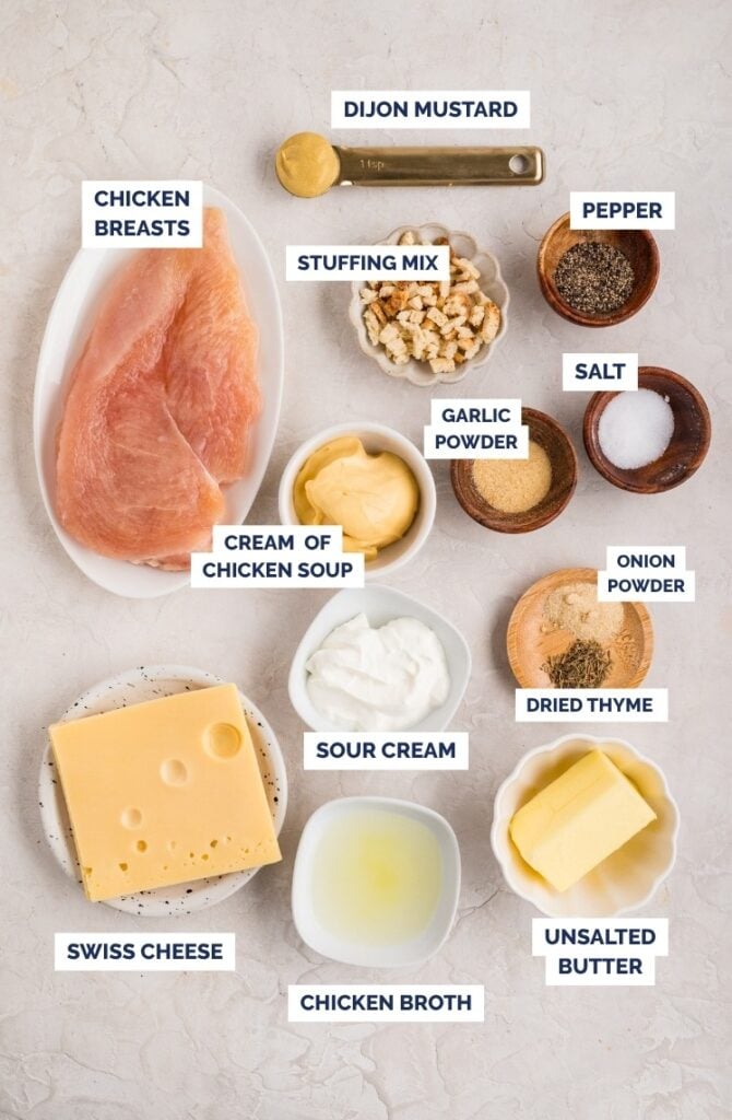 Ingredients for a chicken dish arranged on a countertop, including chicken breasts, stuffing mix, Swiss cheese, sour cream, unsalted butter, chicken broth, cream of chicken soup, Dijon mustard, and various seasonings.