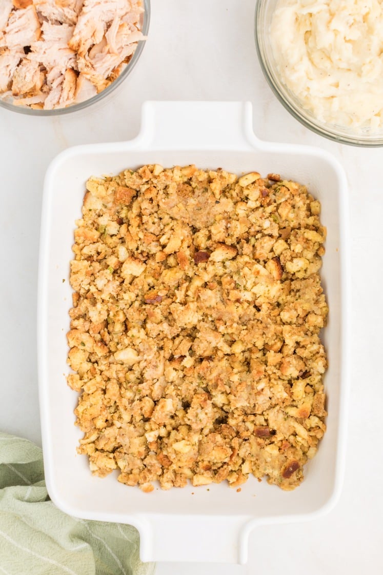 A white baking dish filled with golden-brown stuffing sits on a light surface, with bowls of shredded turkey and mashed potatoes nearby. A green cloth is partially visible in the lower left corner.