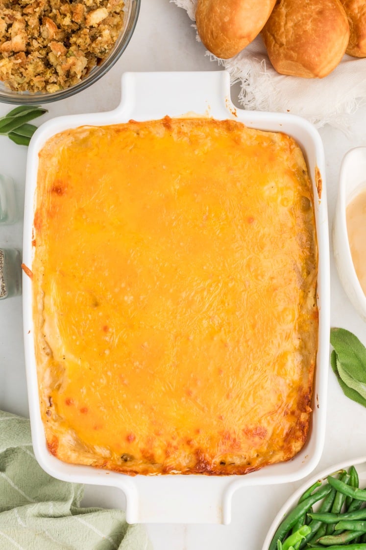 A baked casserole in a white dish, topped with melted golden cheese, surrounded by bowls of stuffing, green beans, and bread rolls on a white surface.