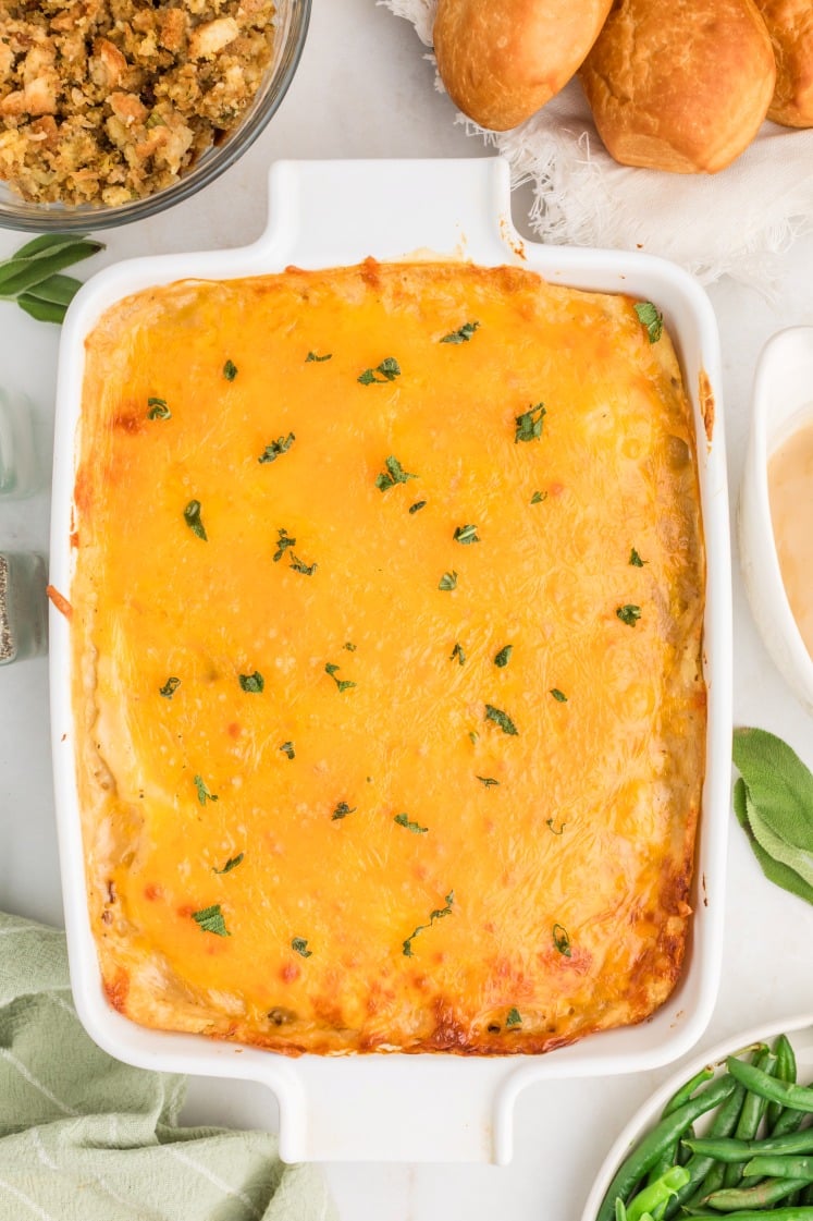 A baked casserole topped with melted cheddar cheese and sprinkled with chopped herbs, in a white rectangular dish, surrounded by rolls, green beans, and stuffing on a marble surface.