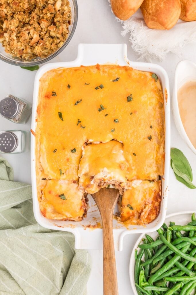 A casserole dish filled with a cheesy baked casserole, with a wooden spoon lifting a portion. Surrounding the dish are green beans, stuffing, dinner rolls, and a beige sauce. A green striped towel is nearby.