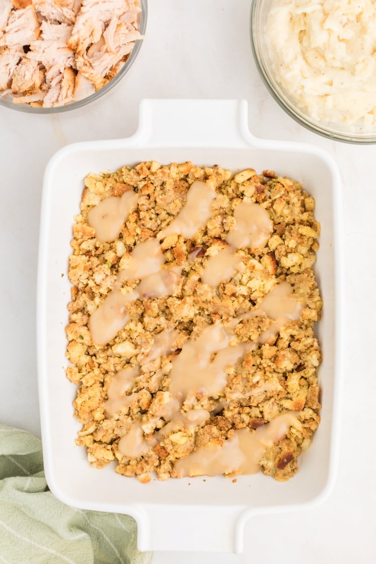 A white baking dish filled with stuffing topped with brown gravy sits on a white surface. Nearby are bowls of shredded turkey and mashed potatoes, and a folded green cloth.