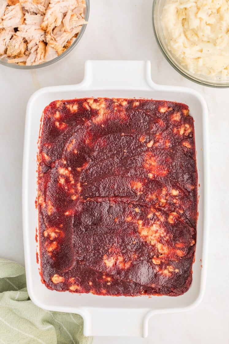 A white baking dish filled with a layer of cranberry sauce over stuffing, next to bowls of shredded turkey and mashed potatoes on a light countertop.