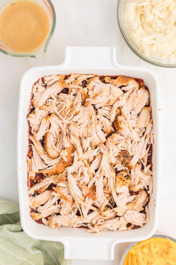 A white baking dish filled with shredded cooked chicken on a layer of stuffing. Surrounding the dish are a bowl of mashed potatoes, a bowl of shredded cheddar cheese, and a measuring cup of gravy.