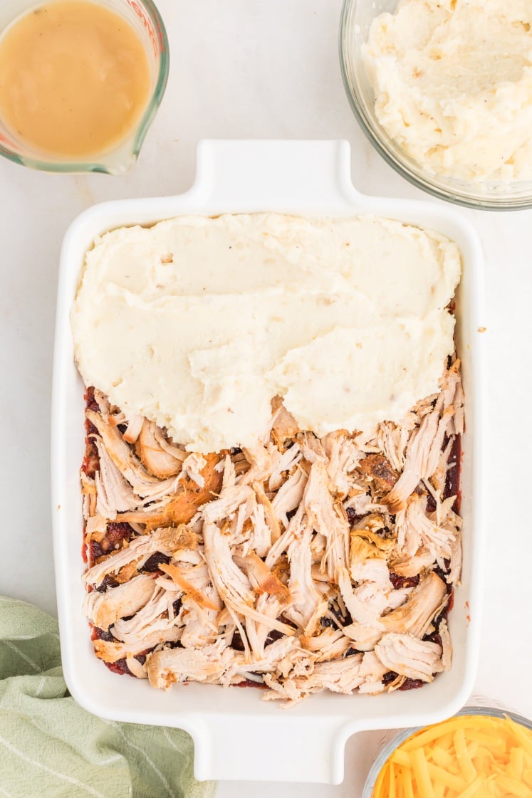 A white baking dish is half topped with mashed potatoes and half with shredded chicken over a red sauce. Surrounding the dish are bowls of grated cheese, mashed potatoes, and a measuring cup of gravy.