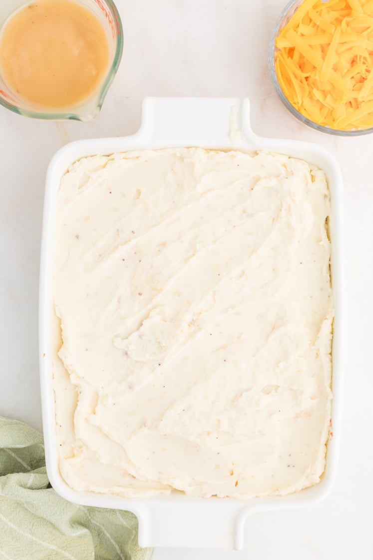 A white baking dish filled with a smooth layer of mashed potatoes sits on a white surface. Nearby are a glass measuring cup with gravy and a bowl of shredded cheddar cheese. A green cloth is partially visible.