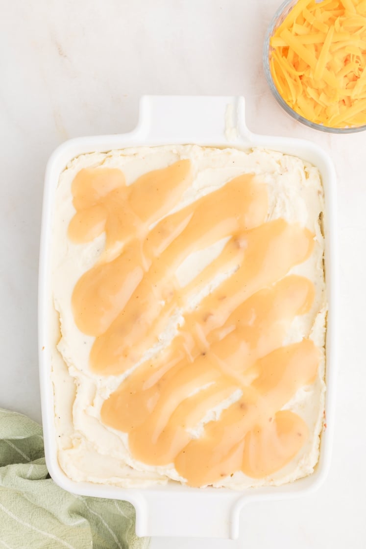 A white baking dish filled with a creamy mashed potato layer topped with light brown gravy. A small bowl of shredded cheddar cheese sits beside the dish on a white surface.