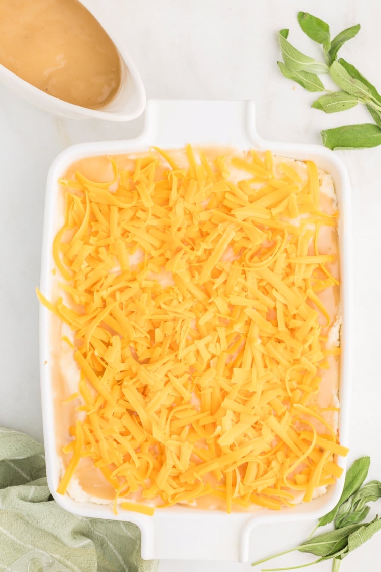 A white baking dish filled with shredded cheddar cheese over a creamy layer, surrounded by green herbs, a light green cloth, and a small white pitcher of gravy on a white surface.