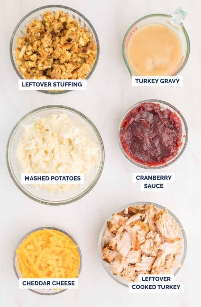 Six bowls on a white surface containing leftover stuffing, turkey gravy, mashed potatoes, cranberry sauce, cheddar cheese, and cooked turkey, each labeled with text.