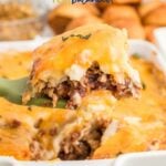 A large serving of cheesy Thanksgiving leftovers casserole is being lifted from a white baking dish; bread rolls and more food are in the background. Text above reads: “Thanksgiving Leftovers Casserole.”.