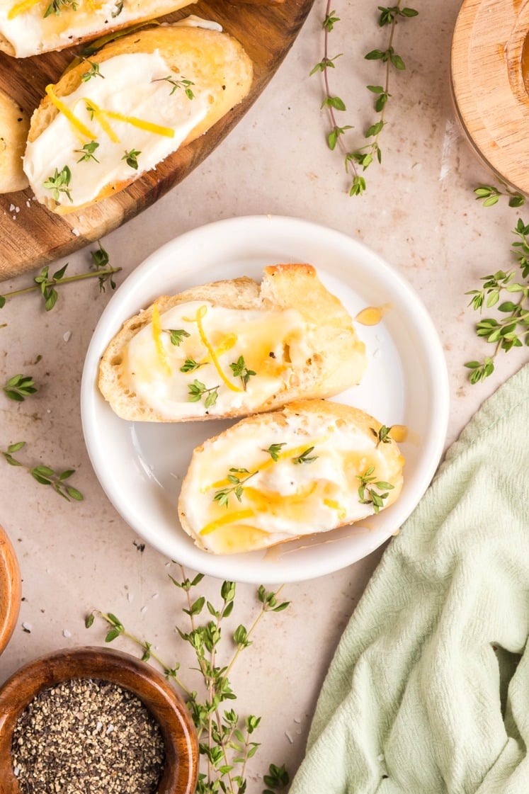 Two slices of bread with creamy cheese, lemon zest, honey, and fresh thyme on a white plate, surrounded by thyme sprigs, a pepper bowl, and a green cloth on a light countertop.