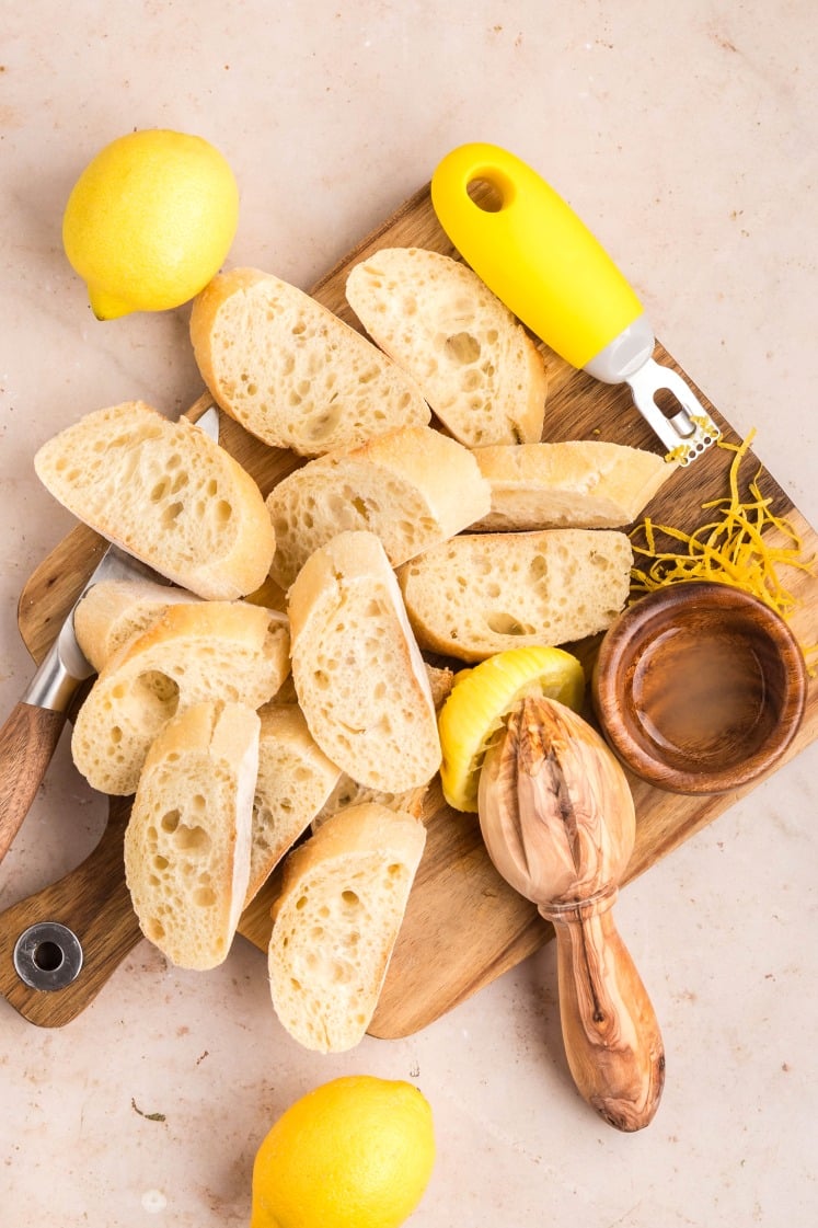 Sliced baguette pieces on a wooden cutting board with a knife, lemons, a zester, a citrus reamer, lemon zest, and a small wooden bowl on a light countertop.