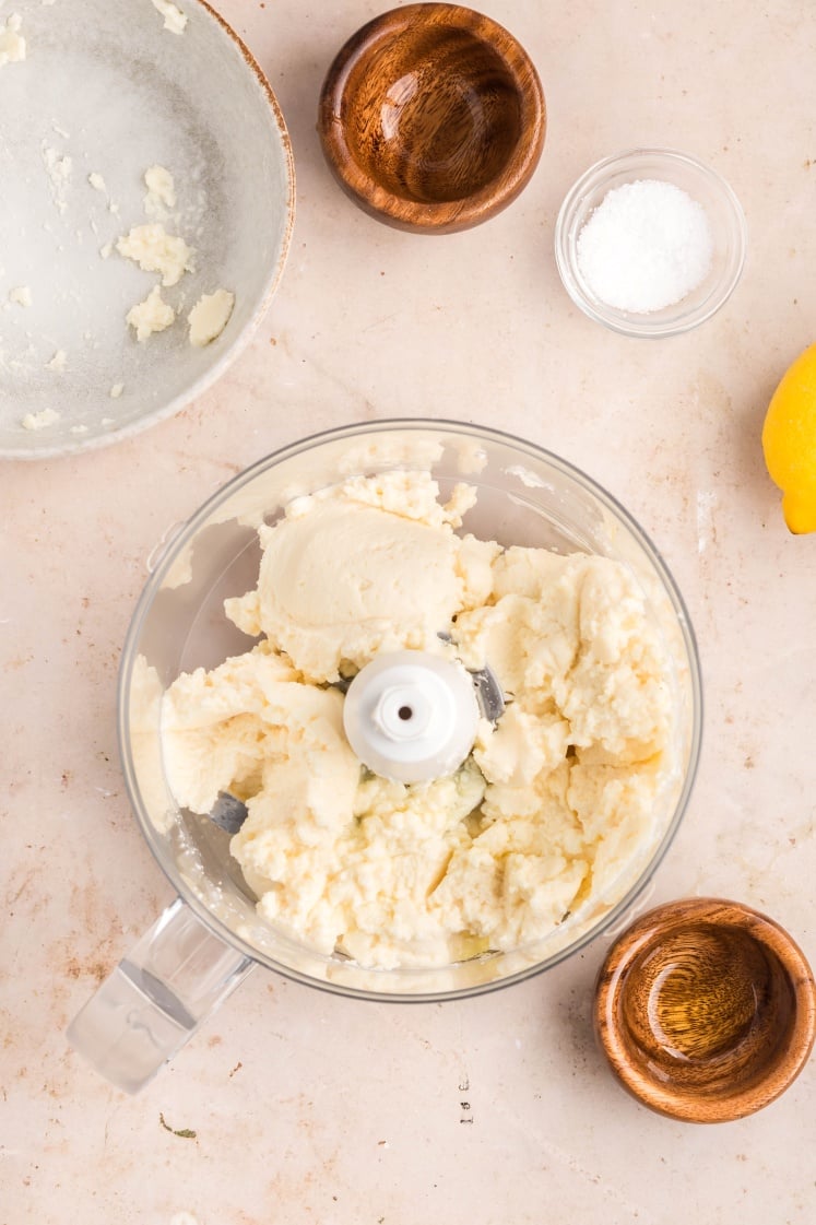 A food processor bowl filled with partially blended butter sits on a countertop, surrounded by small wooden bowls, a lemon wedge, a glass bowl of salt, and a plate with butter remnants.