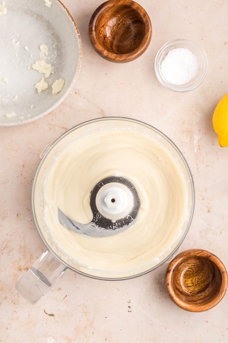 A food processor with creamy white sauce inside sits on a light countertop, surrounded by a bowl with remnants of the mixture, two small wooden bowls, a glass bowl of salt, and part of a lemon.