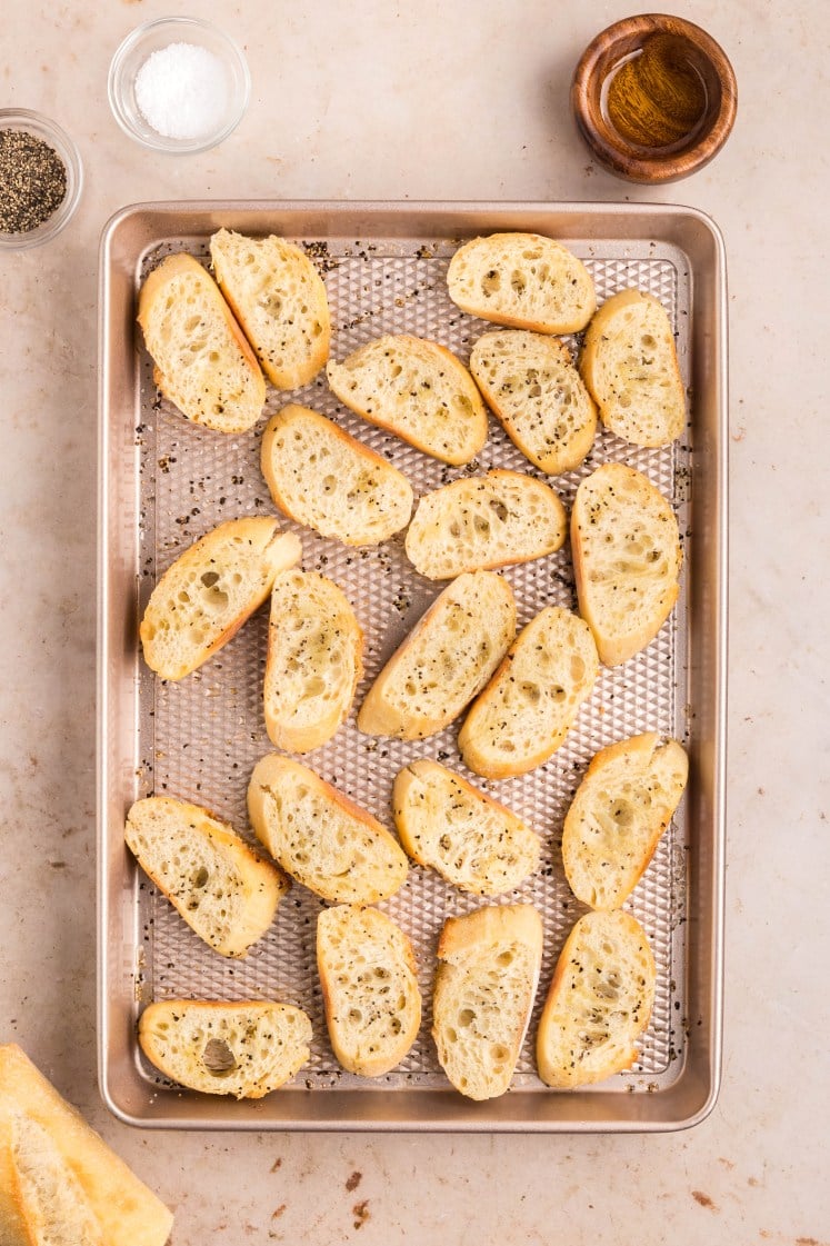 A baking tray with slices of toasted baguette, seasoned with pepper and salt. Salt, pepper, and a small wooden bowl of oil are placed nearby on a light-colored surface.