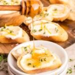 A honey dipper drizzles honey over toasted bread topped with creamy cheese, fresh thyme, and lemon zest. More prepared toasts are visible in the background on a wooden board.