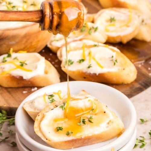 A honey dipper drizzles honey over toasted bread topped with creamy cheese, fresh thyme, and lemon zest. More prepared toasts are visible in the background on a wooden board.