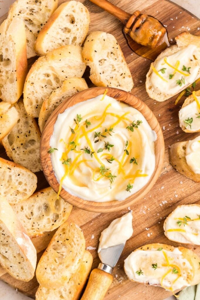 A wooden board with sliced baguette, a bowl of creamy dip drizzled with honey and garnished with fresh herbs, a honey dipper, and a spreading knife with dip. Some baguette slices are spread with dip and herbs.