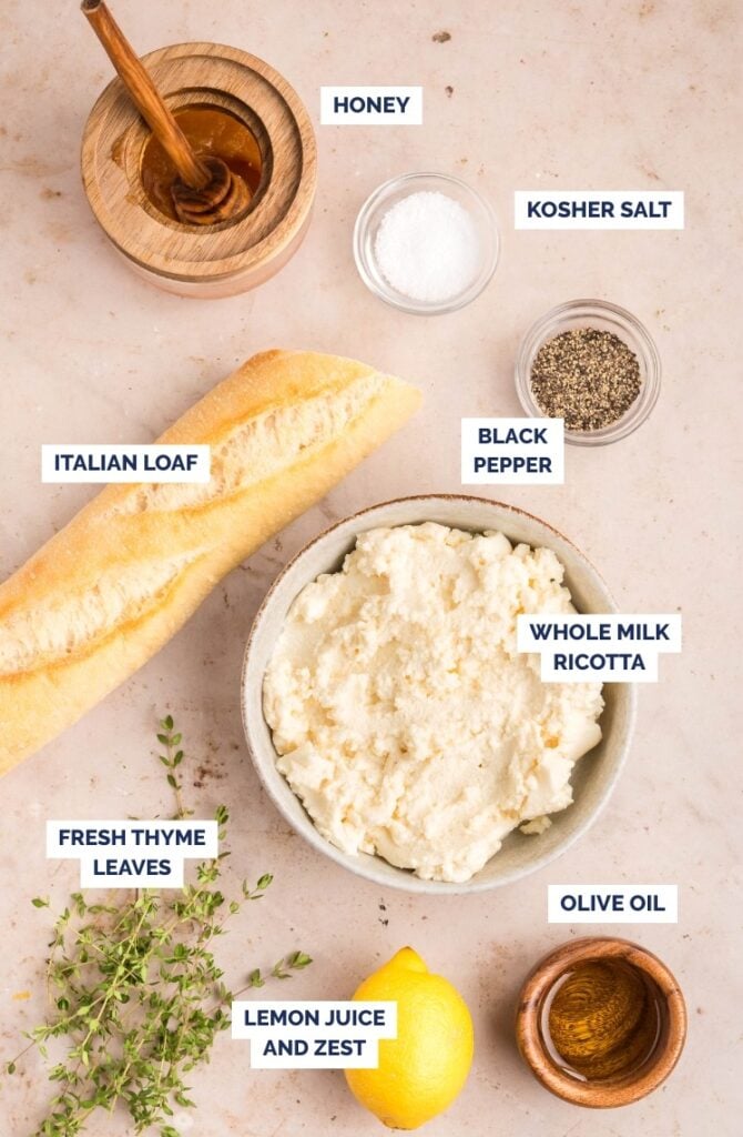 A flat lay of ingredients labeled: honey in a jar, kosher salt, black pepper, whole milk ricotta in a bowl, Italian loaf, fresh thyme leaves, lemon juice and zest, and olive oil on a light surface.