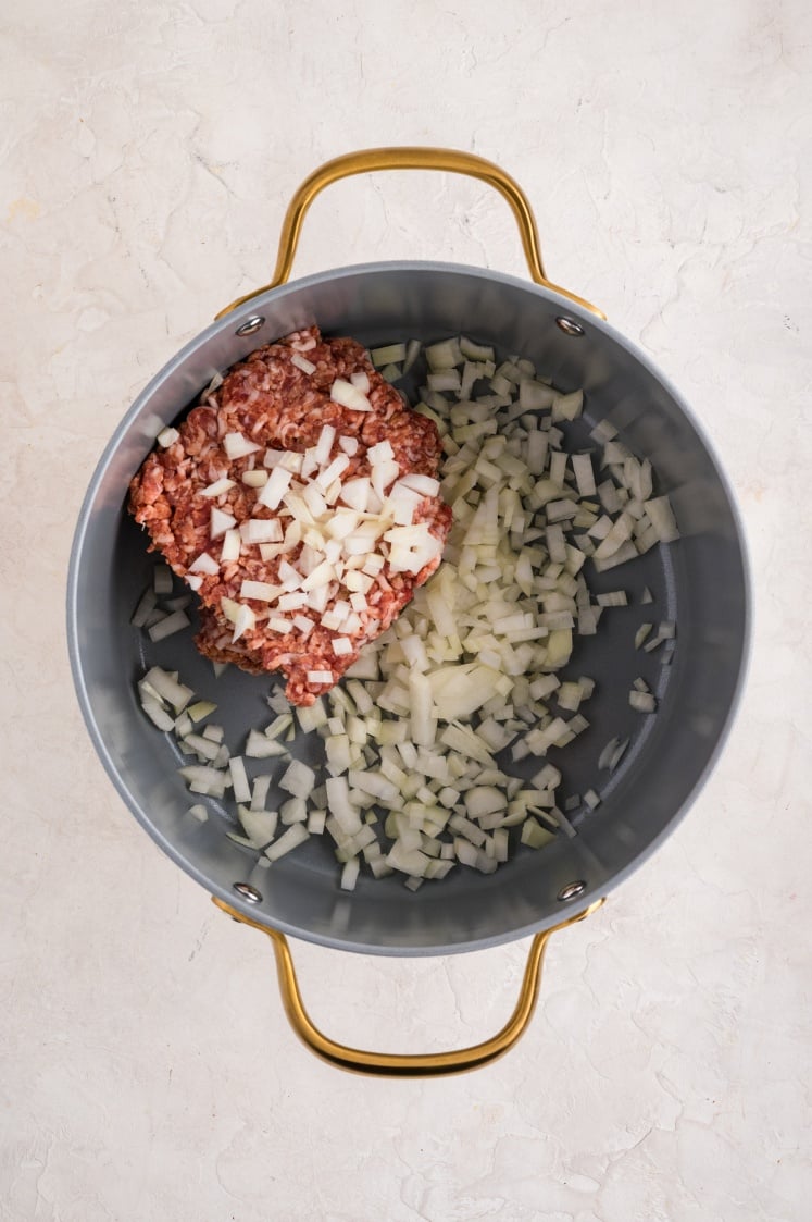 Ground beef and chopped onions in a large gray pot with gold handles, perfect for starting a flavorful Kale and White Bean Sausage Soup, placed on a light-colored textured surface.