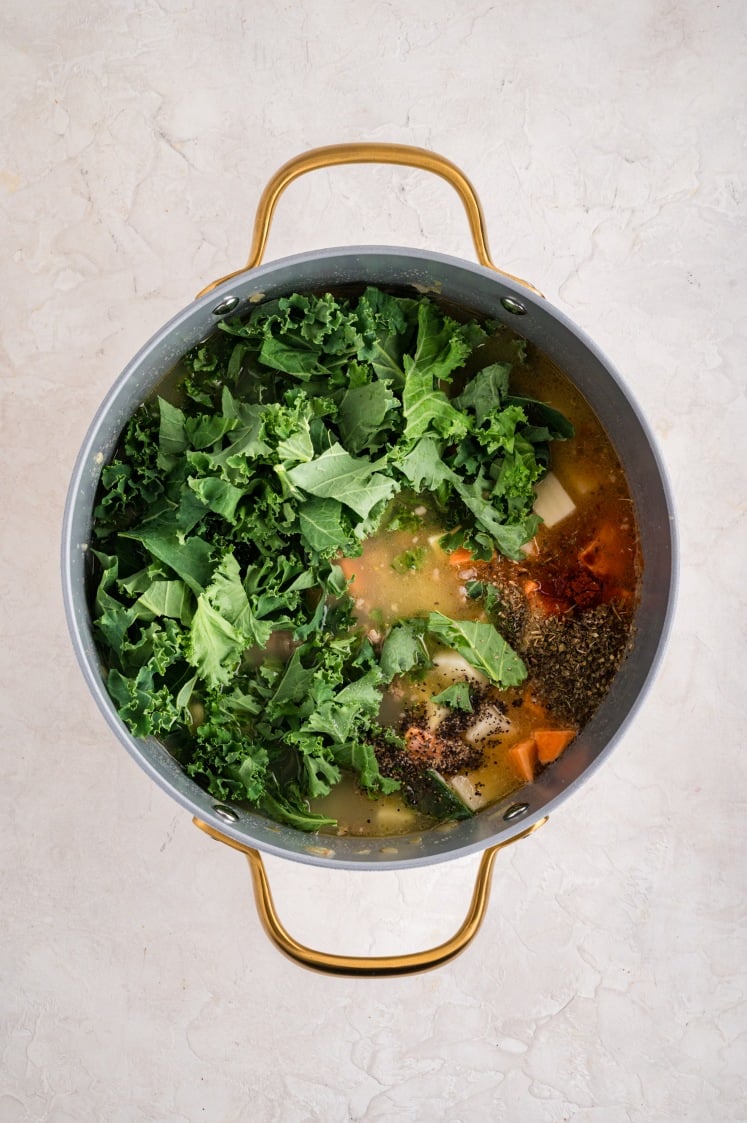 A large pot with gold handles contains Kale and White Bean Sausage Soup ingredients, including fresh kale leaves, chopped vegetables, assorted spices, and broth, ready to be cooked. The pot is placed on a light-colored surface.