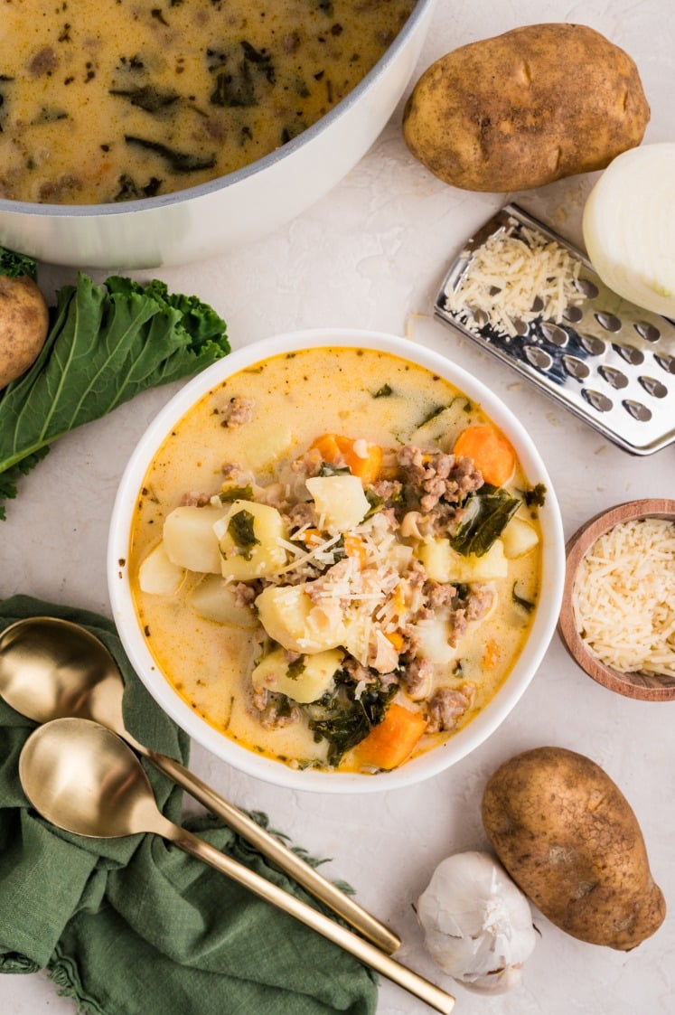 A bowl of creamy Kale and White Bean Sausage Soup with potatoes, sweet potatoes, kale, and grated cheese is surrounded by a pot of soup, russet potatoes, onion, shredded cheese, garlic, gold spoons, and a green napkin.