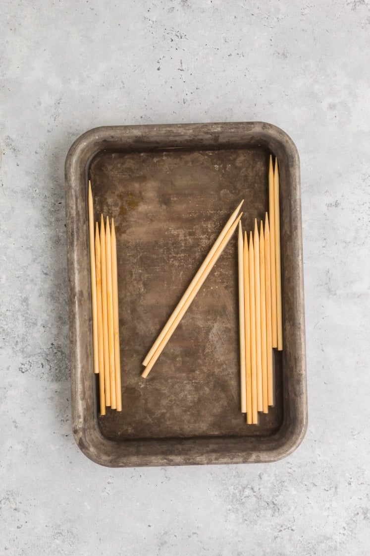 Wooden skewers soaking in a shallow metal tray filled with water, arranged in rows on a gray countertop.
