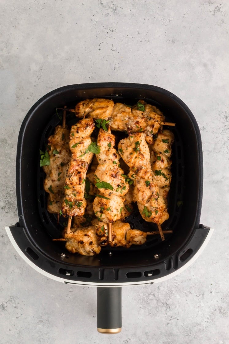 Air fryer basket filled with cooked chicken skewers, browned and sprinkled with fresh parsley, sitting on a light gray countertop.