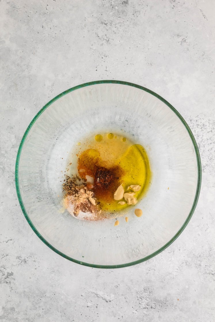Glass bowl with olive oil, spices, Dijon mustard, garlic, and seasonings being mixed.