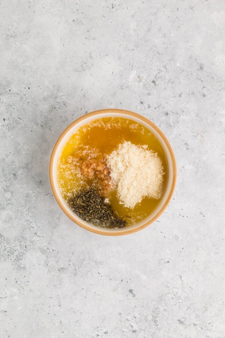 A small bowl filled with melted butter, grated Parmesan cheese, minced garlic, and dried Italian seasoning on a light gray countertop, ready to be mixed for a sauce.