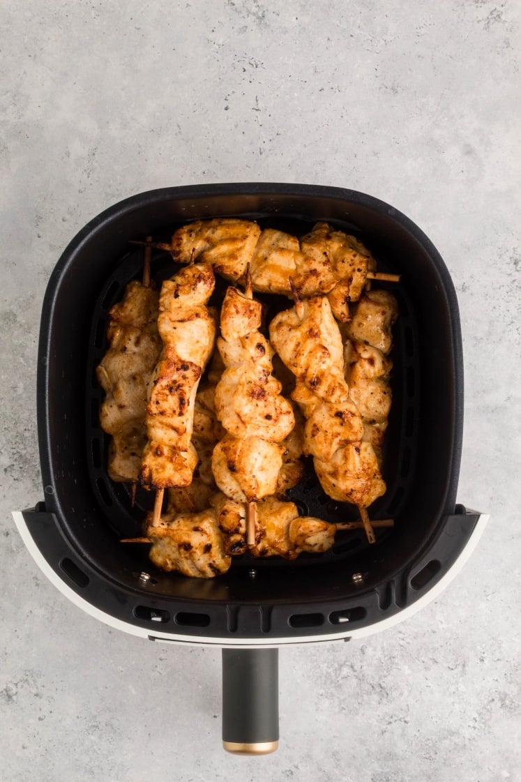 Golden air fryer chicken skewers cooked on wooden sticks inside a black and white air fryer basket, seasoned and lightly charred.