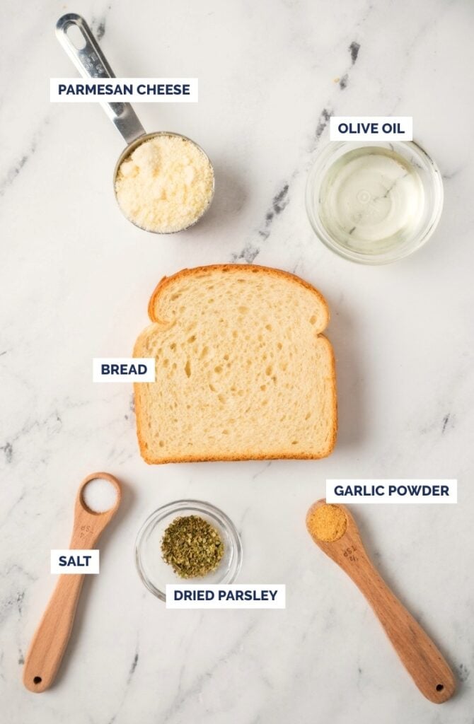 A slice of bread on a marble surface, surrounded by labeled ingredients: a cup of Parmesan cheese, a bowl of olive oil, a spoon of garlic powder, a spoon of salt, and a dish of dried parsley.