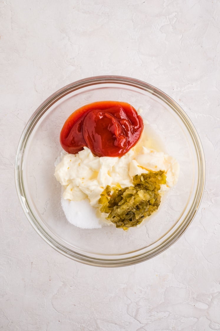 Glass bowl with mayonnaise, ketchup, sweet relish, sugar, and lemon juice being mixed to make sauce.