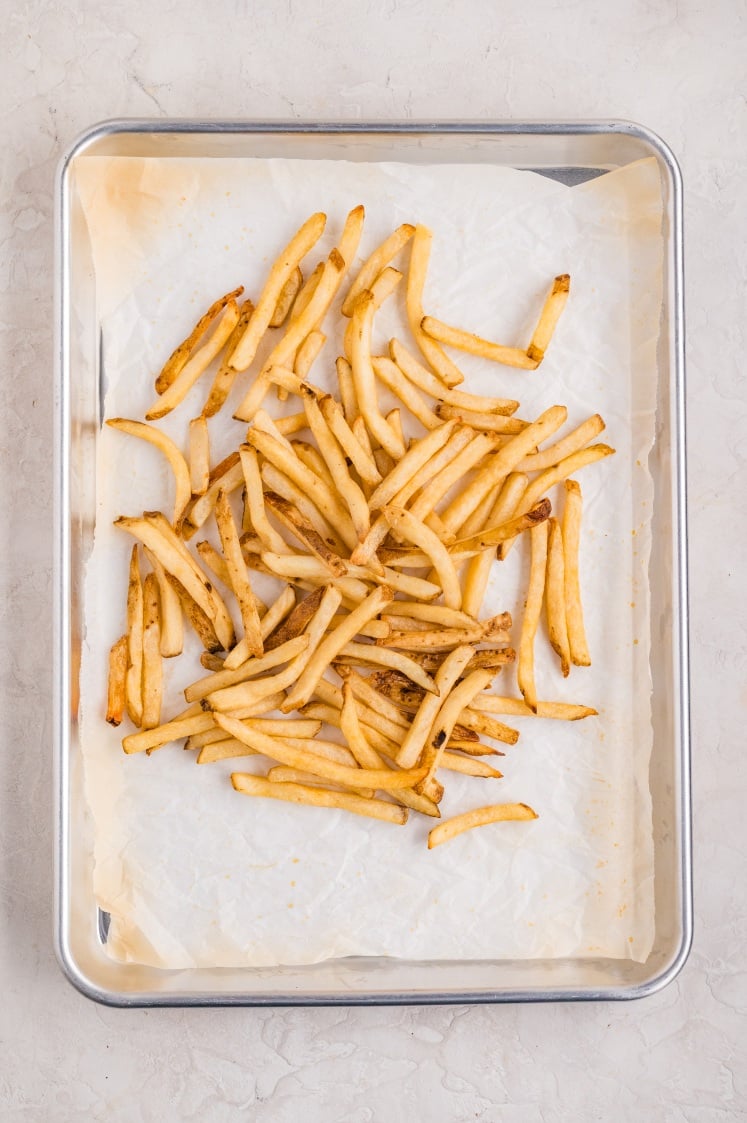 Crispy baked French fries spread on a parchment-lined sheet pan, prepared  with cheese and sauce.