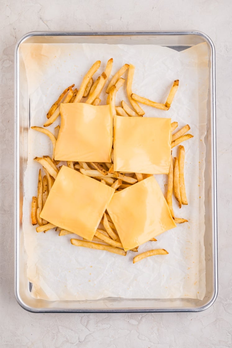 Baking sheet with cooked French fries topped with slices of American cheese, ready to melt.