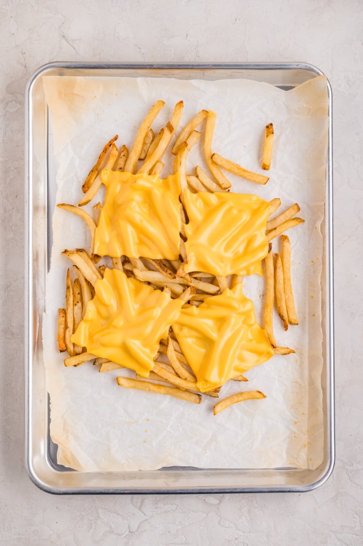 Baking sheet with cooked French fries topped with slices of American cheese, ready to melt