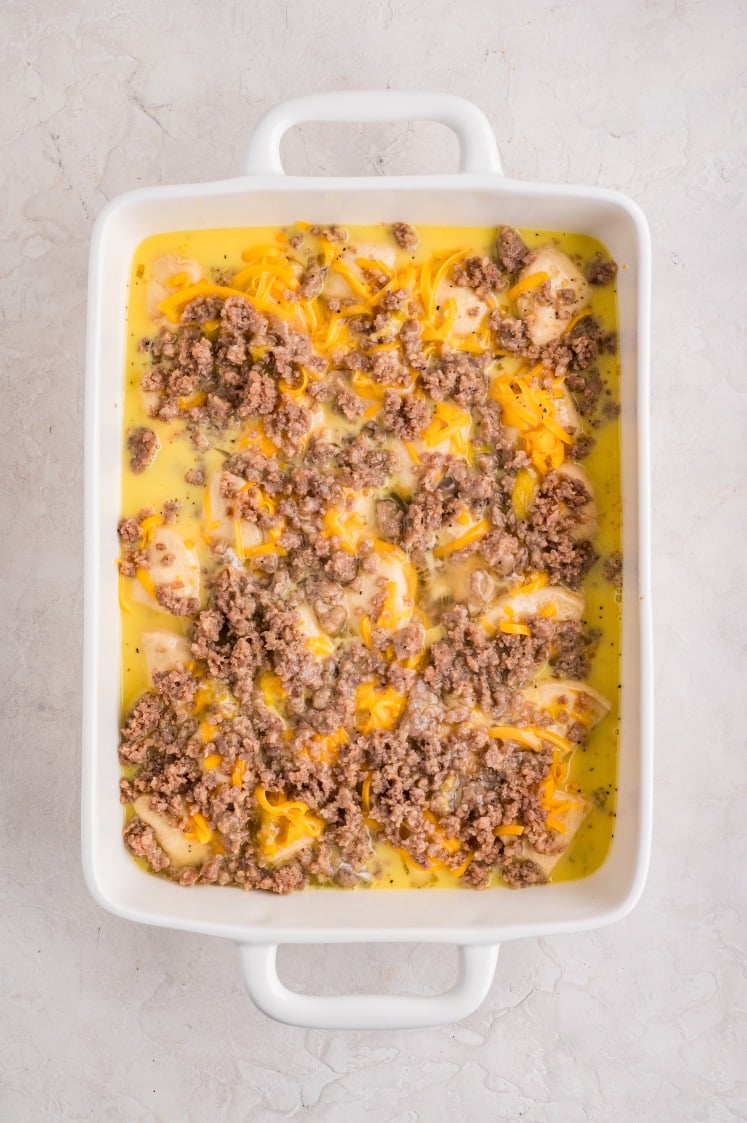 Unbaked biscuits and gravy casserole in a white baking dish, topped with sausage, cheese, and poured egg mixture.