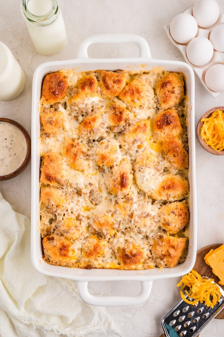 Golden-brown biscuits and gravy casserole fresh out of the oven in a white baking dish, surrounded by eggs, milk, gravy, and shredded cheese.