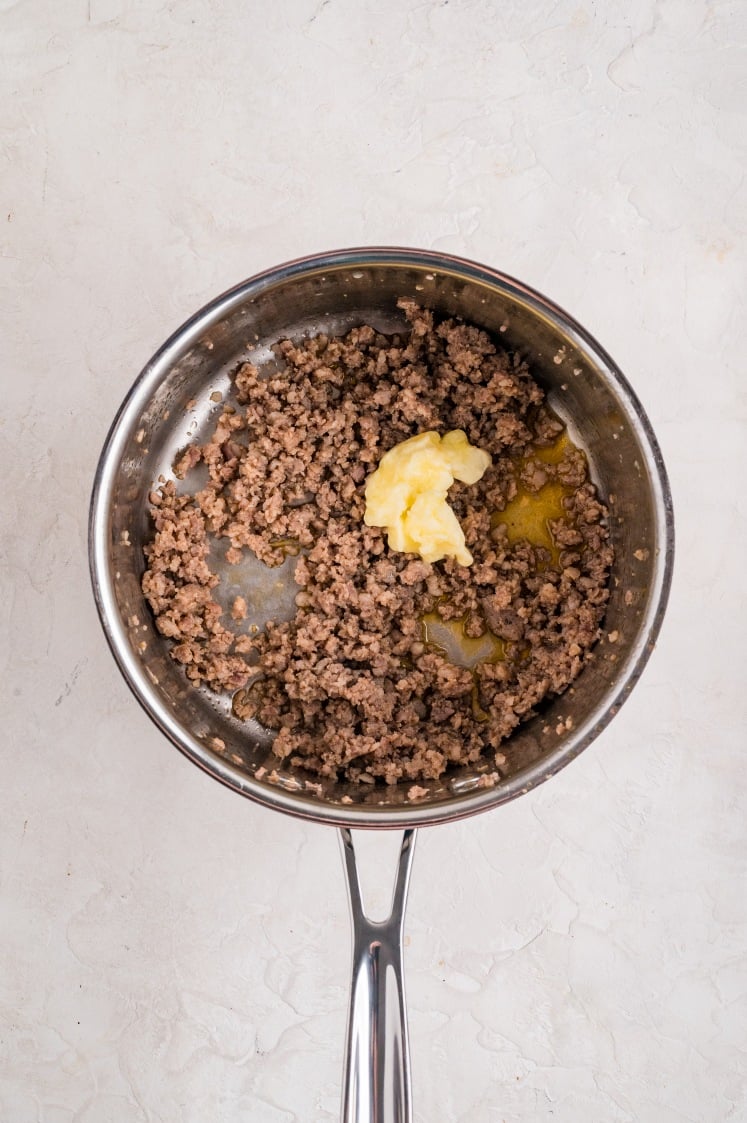 Cooked ground sausage in a stainless-steel skillet with a scoop of butter melting into the meat.