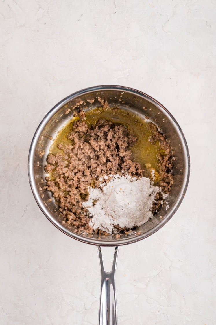 Cooked breakfast sausage in a skillet with melted butter, topped with a scoop of flour to begin making gravy for a biscuits and gravy casserole.