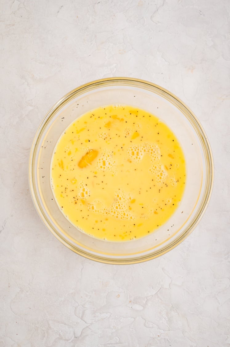 A glass bowl filled with whisked eggs, milk, salt, and pepper, ready to be poured over the casserole.