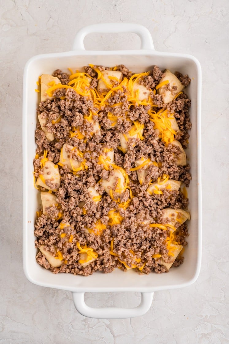 Biscuit pieces layered in a white casserole dish and topped with cooked sausage and shredded cheddar cheese.