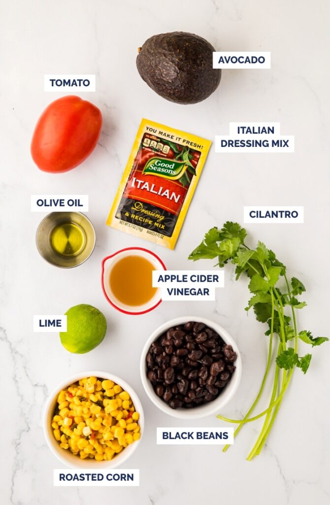 Ingredients on a white surface: a tomato, avocado, Italian dressing mix packet, olive oil, cilantro, apple cider vinegar, lime, black beans, and a bowl of roasted corn. Each item is labeled.